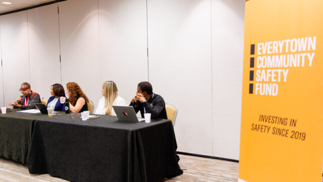 Five panelists sit at a table with a black tablecloth; to their right is an orange placard that reads 