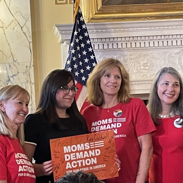 4 women pose for a photo during a bill signing. Three wear red; one wears a black blazer and holds a red 