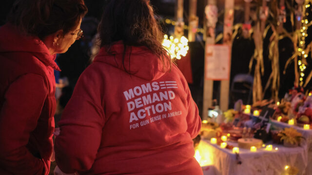 Two Moms Demand Action volunteers in red hooded sweatshirts stand with their backs to the camera as they stand at a Day of the Dead and gun violence prevention event. An ofrenda with string lights is visible in the background.