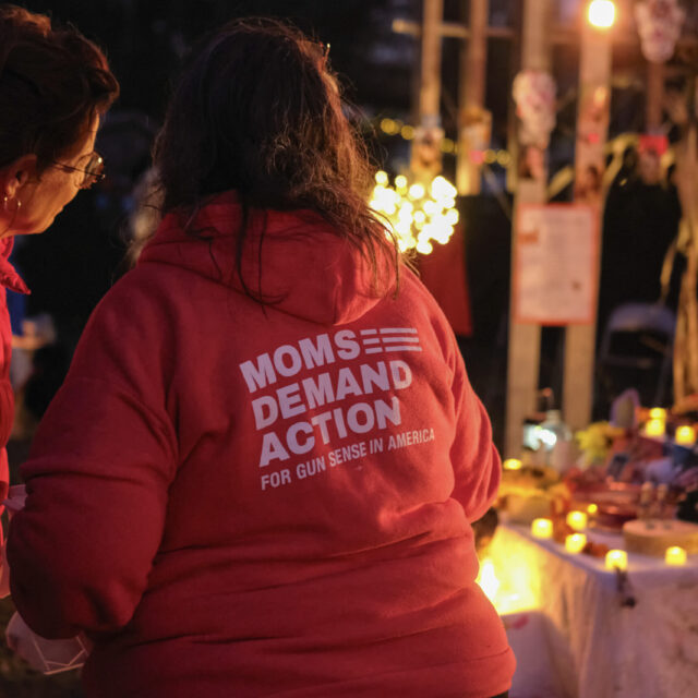 Two Moms Demand Action volunteers in red hooded sweatshirts stand with their backs to the camera as they stand at a Day of the Dead and gun violence prevention event. An ofrenda with string lights is visible in the background.