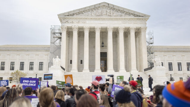 Gun violence prevention advocates rally outside of the Supreme Court. The bottom third of the image shows people in winter clothing—hats, sweatshirts—holding signs that read 