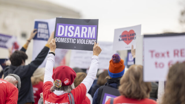 A view from the back of a crowd of Moms Demand Action volunteers protesting in front of the Supreme Court. One volunteer holds a sign that reads 