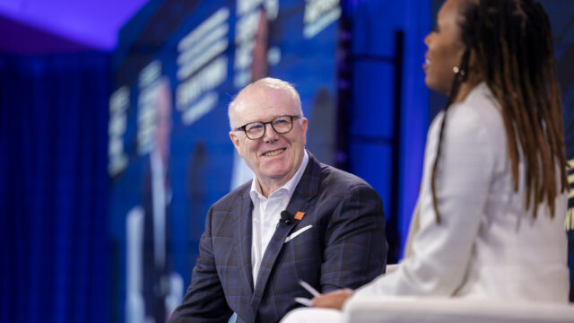 John Feinblatt, president of Everytown for Gun Safety Action Fund, wears a navy and black plaid suit, dark glasses, and a white collared shirt while speaking on a stage with Angela Ferrell-Zabala, executive director of Moms Demand Action.