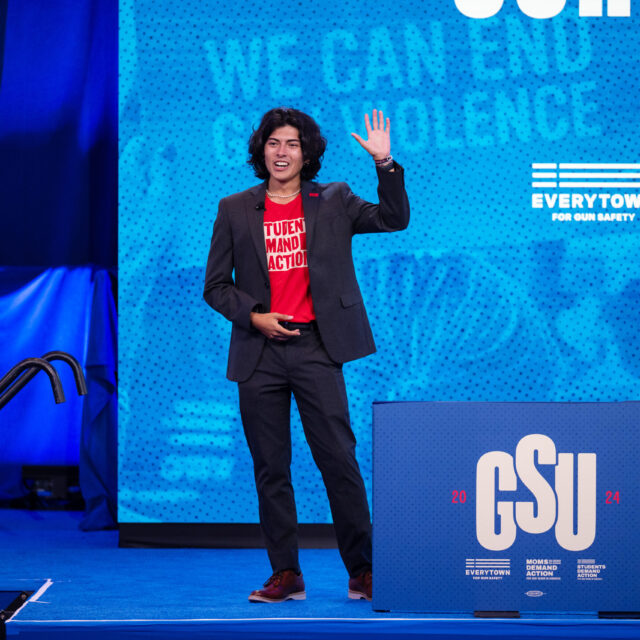 Andres Cubillos has curly dark hair cut just above his shoulders. He is standing against a blue background and wears a black suit with a red students demand action t-shirt. His left hand is raised in a wave.