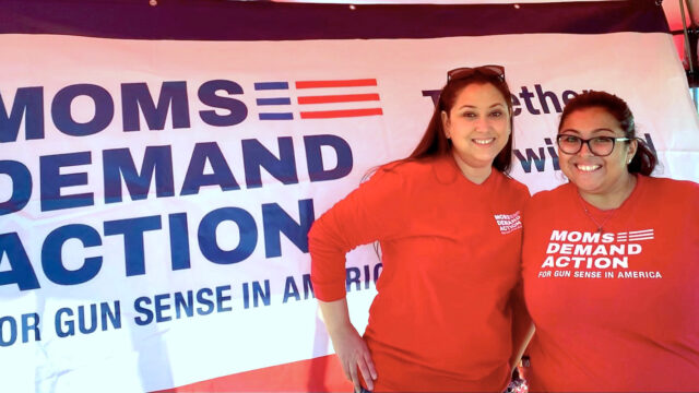 Selina Saenz posing for a photo with another Moms Demand Action volunteer in front of a Moms Demand Action banner