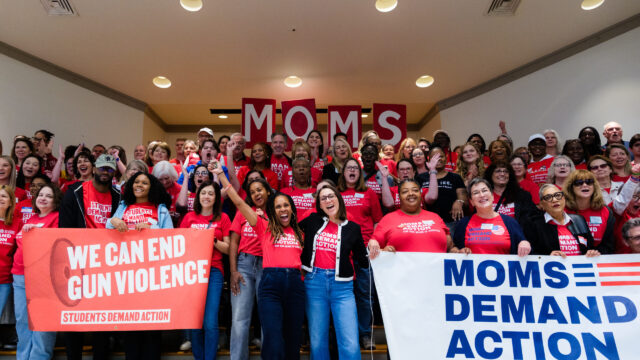 A large group of people, all wearing red Moms Demand Action or Students Demand Action t-shirts, are smiling, clapping, and cheering in a group photo. In the front left, a group of people hold a banner that says 