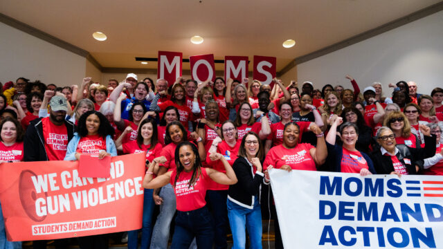 A group of over 100 Moms Demand Action and Students Demand Action volunteers, all wearing red shirts, pose for a photo. Most are holding one arm up and flexing their bicep. Angela Ferrell-Zabala, executive director of Moms Demand Action and Students Demand Action, has her sleeves rolled up and is flexing in between two groups of volunteers who are holding 