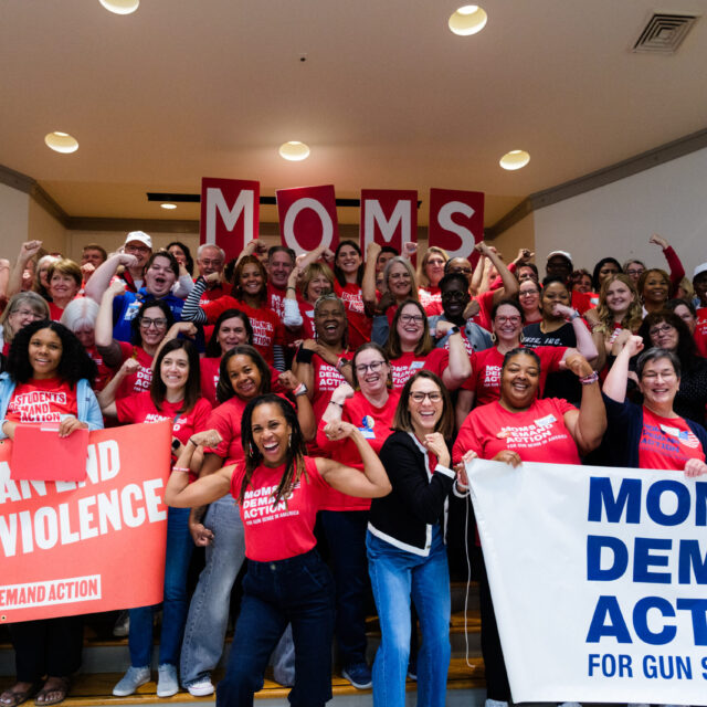 A group of over 100 Moms Demand Action and Students Demand Action volunteers, all wearing red shirts, pose for a photo. Most are holding one arm up and flexing their bicep. Angela Ferrell-Zabala, executive director of Moms Demand Action and Students Demand Action, has her sleeves rolled up and is flexing in between two groups of volunteers who are holding 