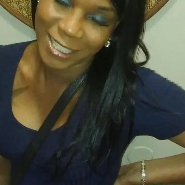 Amber Minor, a Black trans woman, smiles for a photo. Her left hand is propped on her hip, and she is wearing a gold bracelet on that wrist. She wears a navy blue scoop-neck t-shirt and her long black hair is pulled over her left shoulder.