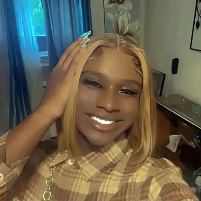 Amiri Jean Reid, a Black trans woman, takes a selfie. She is wearing a brown plaid collared shirt and is holding her right hand against her shoulder-length hair, which is parted down the middle.