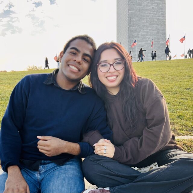 Kyle Clarke Echeverría and Andrea Joseline Rodríguez Ávila pose for a photo while sitting on a sidewalk in front of the Washington Monument. They are smiling and are linking arms.