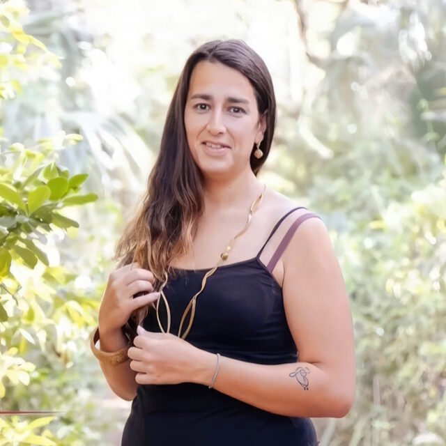 Andrea Doria Dos Passos, a Latina trans woman, poses for a portrait photo. Her long dark hair is parted off to one side and is pushed over her right shoulder. She wears a long, loopy gold necklace and a black tank-top; she has a chunky cork bracelet on her right wrist arm just below her elbow.
