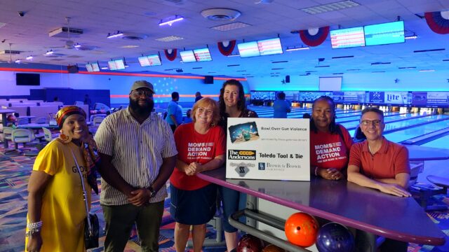 Six people pose for a photo inside of a bowling alley. Two are wearing Moms Demand Action shirts. They all are gathered around a white sign that reads 