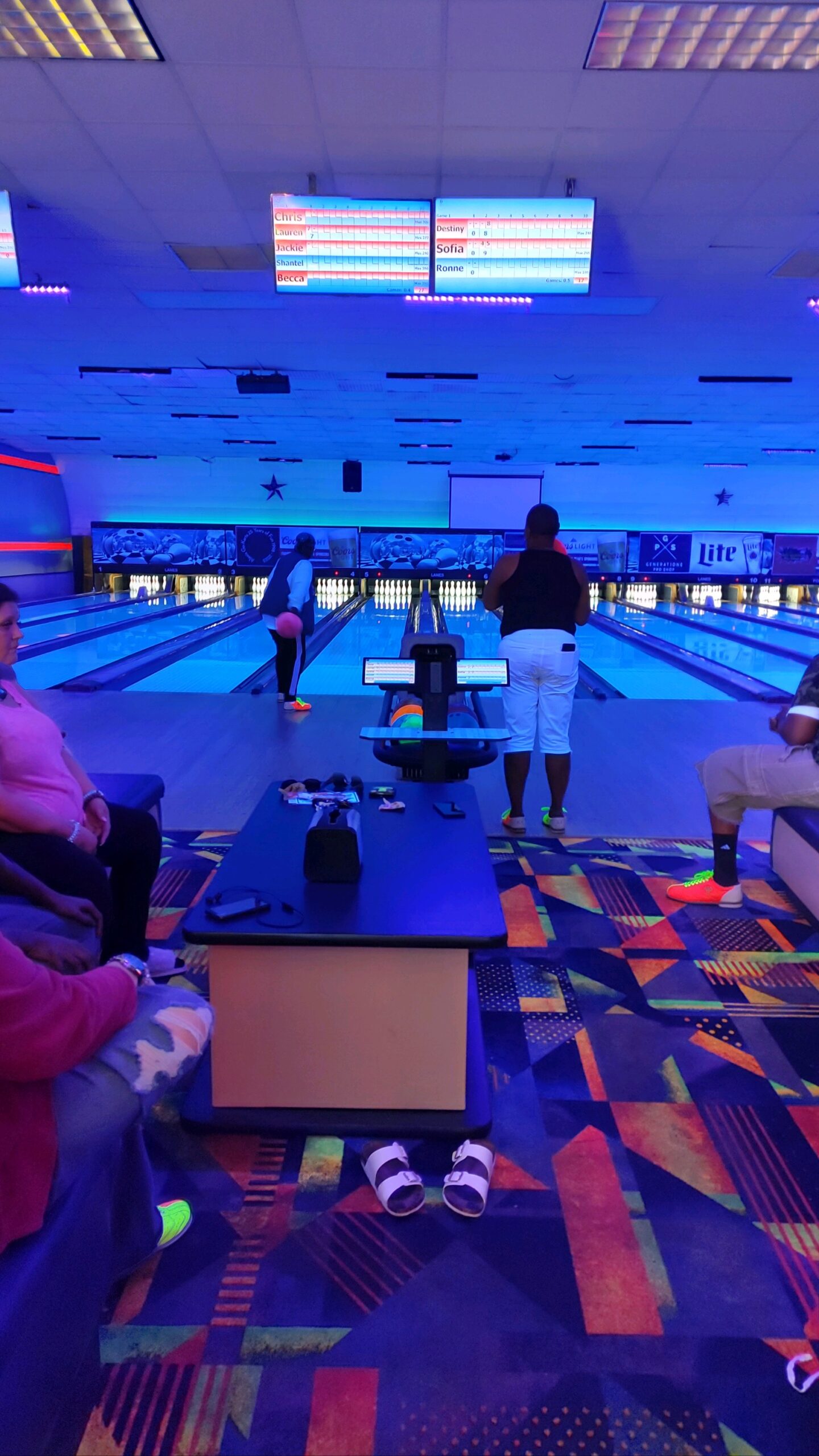 A photo of a woman in white pants and a black tank top (photo is taken from behind) about to bowl a ball down a bowling alley lane.