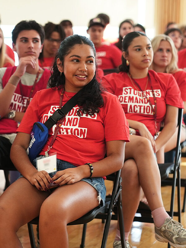 Students Demand Action volunteers sit in rows of folding chairs in an auditorium as they listen to an off-camera speaker. They are all wearing red Students Demand Action t-shirts.
