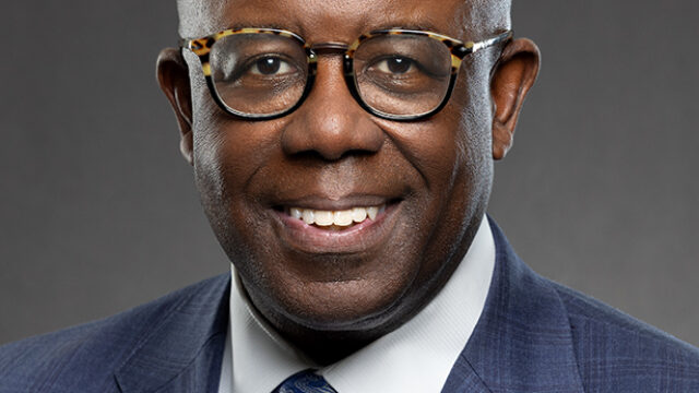 Dr. Barksdale is a Black man with tortoiseshell glasses, a navy blue suit jacket, a white collared shirt, and a blue tie. He is pictured from the upper torso and up in a professional headshot and is smiling, facing the camera, against a dark grey background.