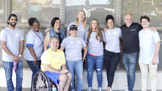 Organizers of the Los Angeles Adaptive Sports Fair pose for a photo and smile. There are 10 people in total, 9 of whom are standing in a row and one of whom is sitting in a wheelchair.