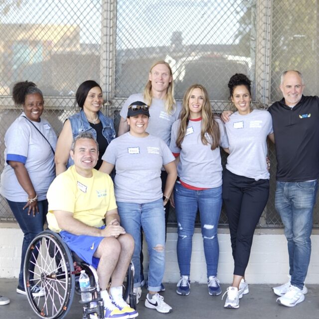 Organizers of the Los Angeles Adaptive Sports Fair pose for a photo and smile. There are 10 people in total, 9 of whom are standing in a row and one of whom is sitting in a wheelchair.