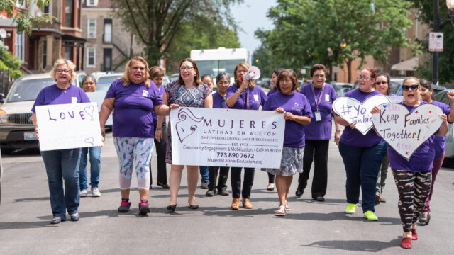 A group of about 15 people march while wearing purple shirts and while holding Mujeres Latinas en Accion signs.