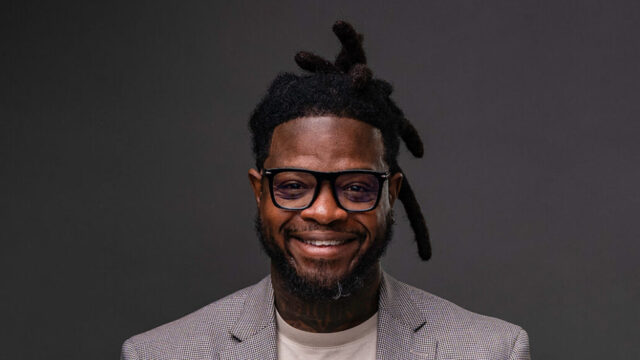 A portrait photo of DuJuan Kennedy, who is posing in front of a professional charcoal-gray background. He wears glasses with a thick black frame, a cream-colored t-shirt, and a thin plaid-stripe blazer.