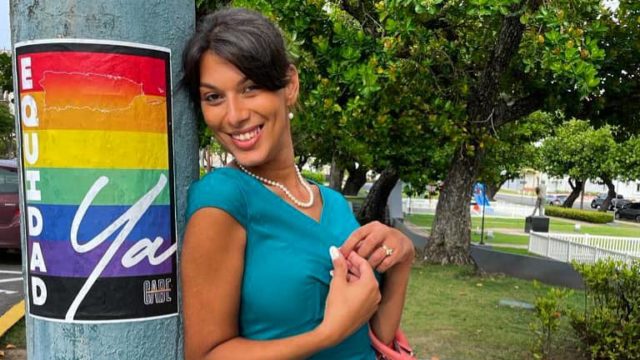 Joanna Cifredo, a human rights activist, smiles while leaning on a pole that has a poster with a rainbow pride flag on it