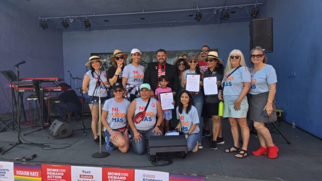 A group of 13 people stand on a stage and pose for a photo. Almost everyone is wearing blue t-shirts or grey tank tops that say 
