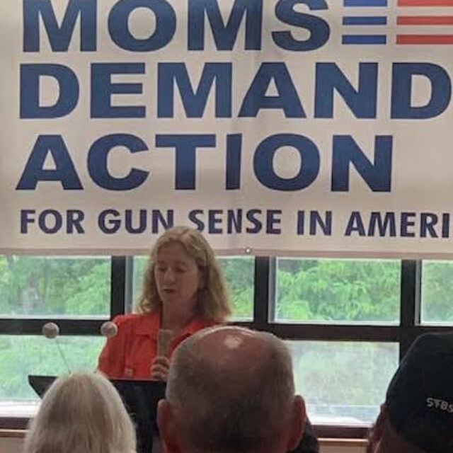 Christine Ilewski, a white woman with shoulder-length blond hair, wears an orange collared shirt and speakers into a microphone. A white banner that reads 