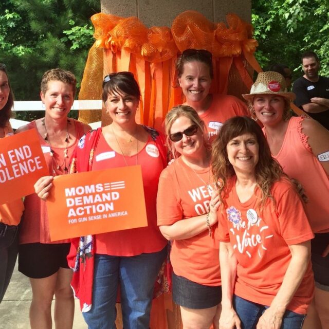 6 women wear orange or red shirts. They smile at the camera one holds an orange sign that reads 