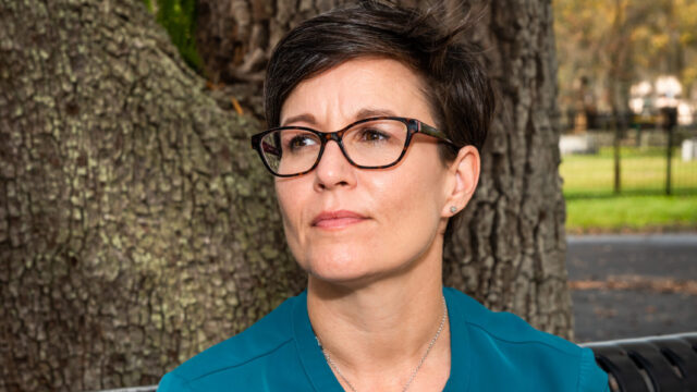 Amanda Goldsmith has short dark hair cropped into a pixie cut. Amanda wears a turquoise blouse and has their left hand crossed over their right wrist. Amanda wears dark glasses and is at a slight profile angle looking to the right of the photo frame.