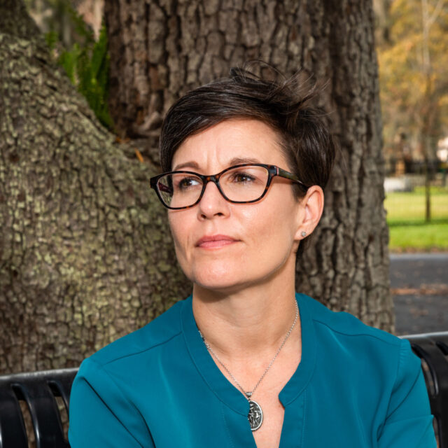 Amanda Goldsmith has short dark hair cropped into a pixie cut. Amanda wears a turquoise blouse and has their left hand crossed over their right wrist. Amanda wears dark glasses and is at a slight profile angle looking to the right of the photo frame.