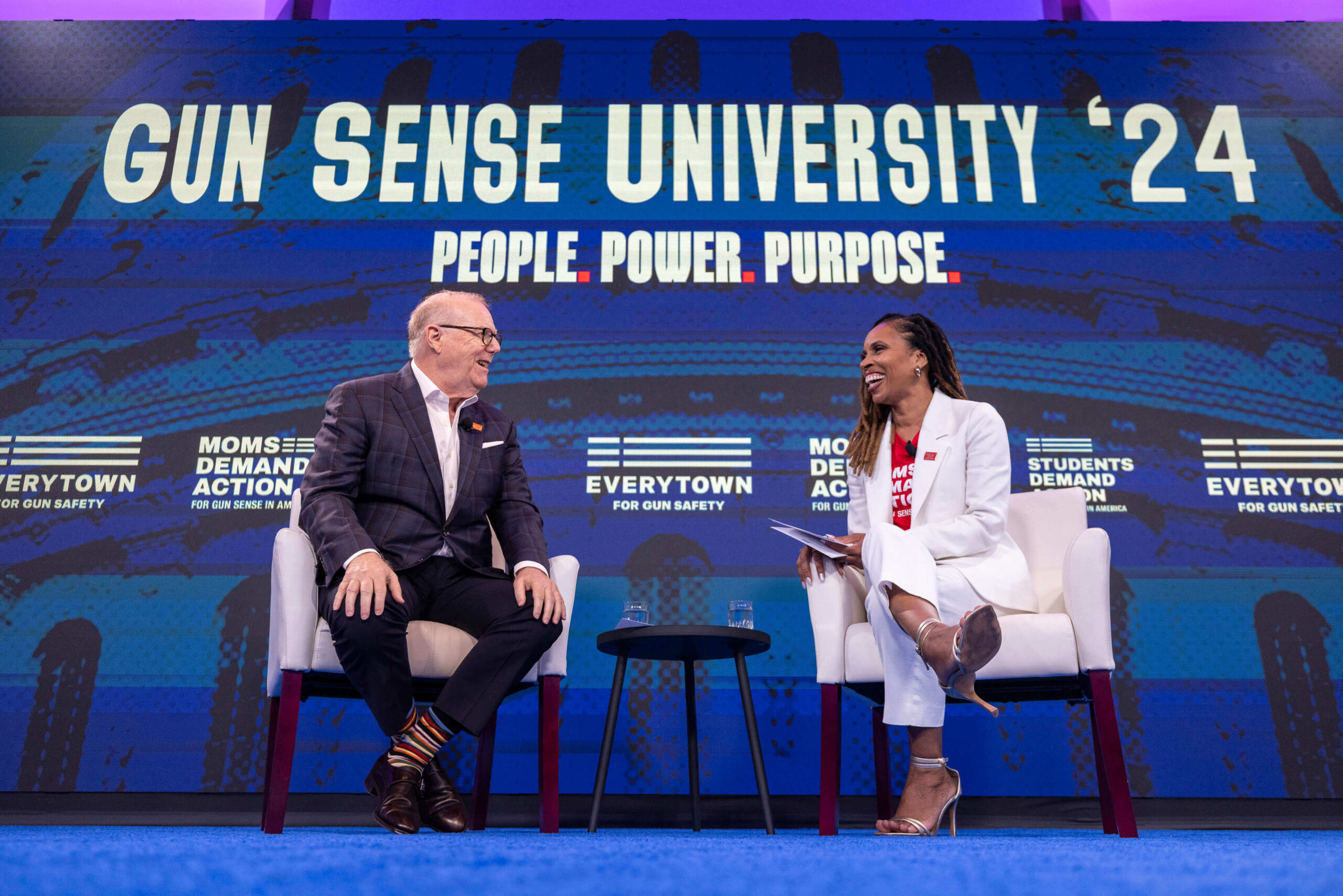 A candid photo of John Feinblatt, a white man in dark pants, a suit jacket, and a white collared shirt, speaking with Angela Ferrell-Zabala, a Black woman wearing a white pantsuit with silver heels and a red Moms Demand Action t-shirt. The two sit on white chairs set side-by-side with a table in between; above them is a sign that reads "Gun Sense University '24: People. Power. Purpose."