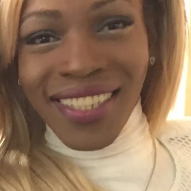 Honee Daniels, a Black trans woman, takes a selfie. Her light blond hair is straight and falls to around her shoulders. She is wearing a white turtleneck top with a mesh white layer on top of the turtleneck. She wears small stud earrings and smiles at the camera.
