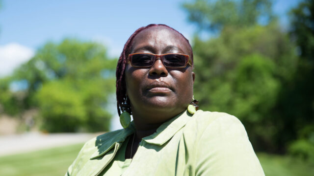 Greta Willis, a Black woman wearing a pastel green collared shirt, matching pastel earrings, and dark, narrow oval sunglasses, turns toward the camera and poses for a photo. Green grass and trees are visible in the background behind her.