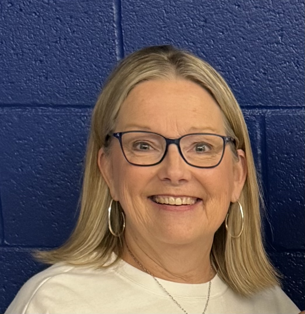 An image of Patty Matthews, a woman with light skin, dark glasses, and a white shirt. She is smiling and her shoulder-length blonde hair is straight and parted down the center. She wears medium-size hoop earrings and a silver necklace chain.