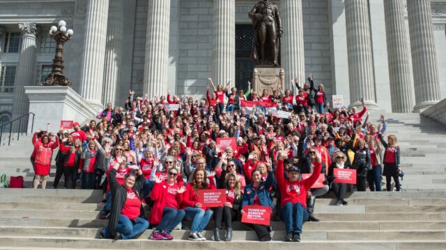 A group of over 50 Moms Demand Action Volunteers cluster on the front steps of a government building, wearing red t-shirts, jeans, and holding Moms Demand Action red signs.
