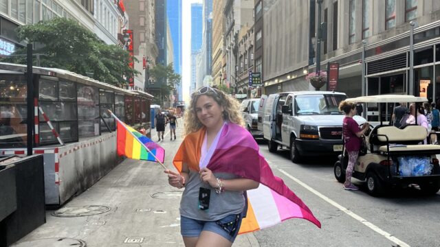 A person with light skin and shoulder-length curly blond hair poses for a photo int he middle of a street. Grey tall buildings are pictured on either side of them and recede into the distance. They have white sunglasses in their hair and hold a rainbow flag in their right hand. They have the lesbian pride flag wrapped around their shoulders. They wear a grey t-shirt, blue denim shorts, and white shoes with a rainbow on the side.