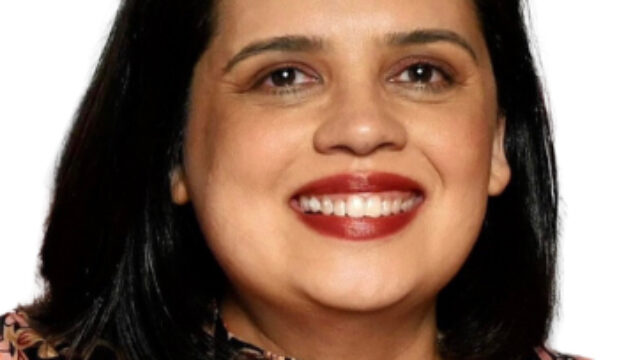 A shoulders-up headshot of Neha Gill, a woman with dark, shoulder-length hair, red lipstick, and wearing a black shirt with red and yellow flowers.