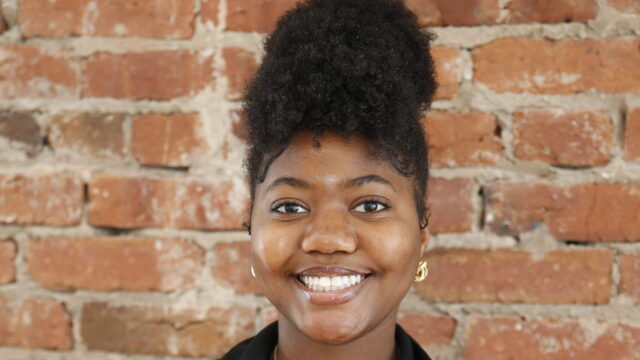 Iyanah Forbes, an HBCU student and the chartering president of Students Demand Action, poses for a professional photo. She stands in front of a brick wall and wears a black blazer, a gold necklace with a pendant, and small dangly gold earrings.