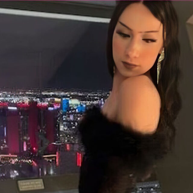 Jazlynn Johnson, a trans woman, poses for a photo in front of a window at night that overlooks a city skyline. Jazlynn wears a black off-the-shoulder top that appears to have a fluffy top hem; her dark hair is parted in the middle and is in loose curls down her back. She is looking over her left shoulder at the camera and wears long silver metal earrings.