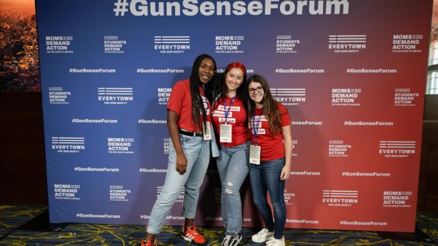 Alanna Miller and two other Students Demand Action volunteers pose for a photo in front of a step and repeat background at the Gun Sense Forum