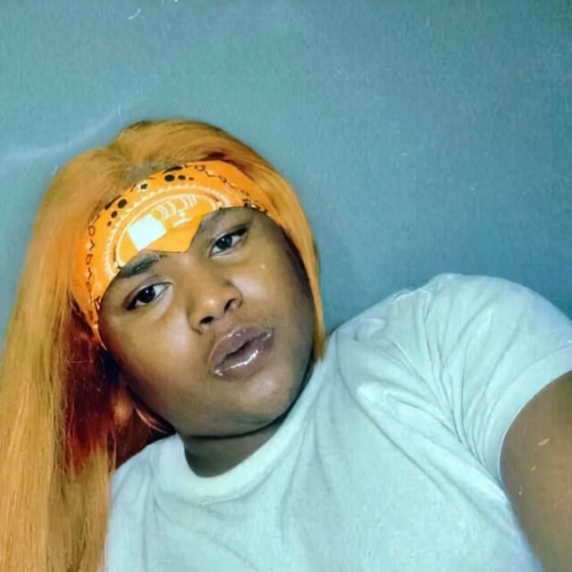 Kejuan Richardson, a Black gender-nonconforming man, poses for a photo. Kejuan is leaning against a teal blue wall; Kejuan has long hair pulled over his right shoulder and held back from his forehead by an orange bandana.