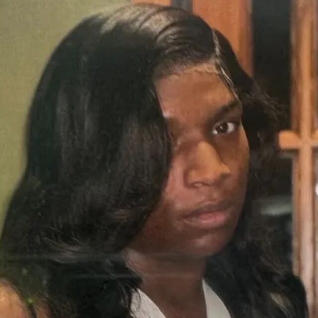 Kenji Spurgeon, a Black trans woman, poses for a photo in front of a green wall and a wooden door with small windows. She wears a white halter top and her black shoulder-length hair is parted down the side; part of the hair falls in front of her right eye.