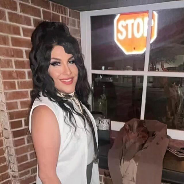 Kitty Monroe, a Latine trans person, poses for a photo in front of a brick wall and a window. Kitty wears a white sleeveless top and a metallic necktie; their long, wavy black hair is pulled into a half-up, half-down style.