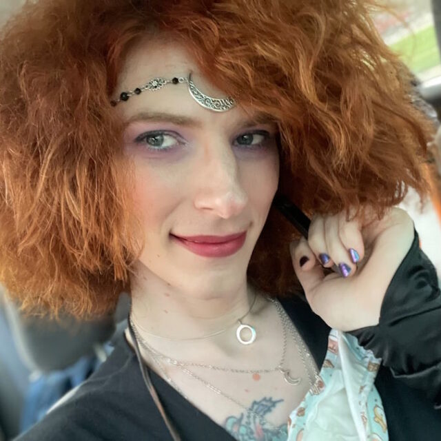 Liara Tsai, a white trans woman, takes a selfie while sitting in a car. Her hair is wavy, red, and cropped to around her chin; she wears red lipstick. She has a metallic moon bangle across her forehead and wears layered necklaces with moon pedants. She wears a black long-sleeve top.