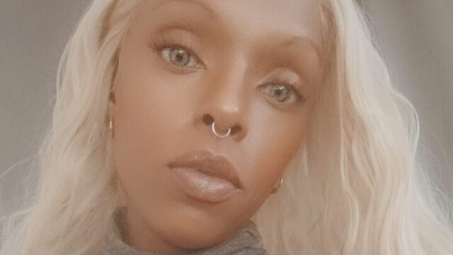 Marquita Wisley, a Black woman, takes a selfie. She is wearing a grey turtleneck sweater and a large aqua blue ring with a silver setting.