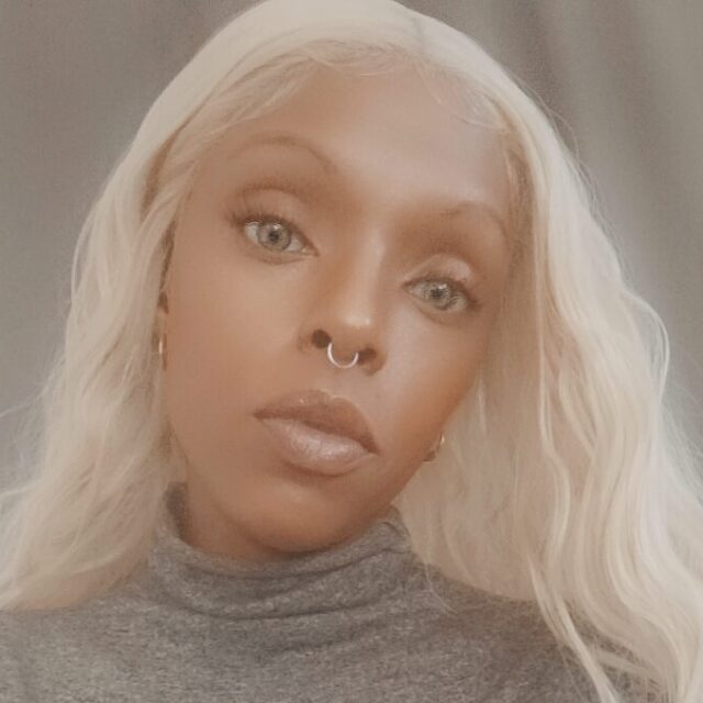 Marquita Wisley, a Black woman, takes a selfie. She is wearing a grey turtleneck sweater and a large aqua blue ring with a silver setting.