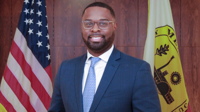 City of Memphis Paul A. Young poses for a professional photo. He is wearing a navy suit jacket, a white collared shirt, and a blue patterned tie. He wears glasses with a light frame and a metal watch. The background is a wood-paneled wall, and he stands between the American flag and the flag of Memphis, Tennessee.