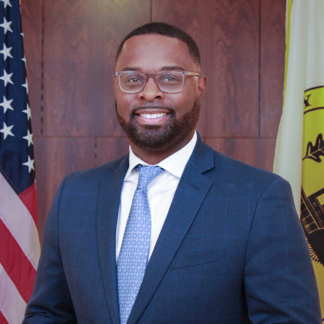 City of Memphis Paul A. Young poses for a professional photo. He is wearing a navy suit jacket, a white collared shirt, and a blue patterned tie. He wears glasses with a light frame and a metal watch. The background is a wood-paneled wall, and he stands between the American flag and the flag of Memphis, Tennessee.