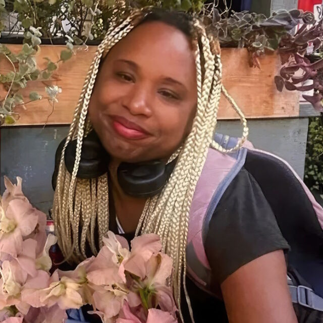 Michelle Henry, a Black trans woman, poses for a photo with pink flowers in front of her and a grey-and-brown planter box behind her. Her hair is plaited in blond braids that reach near her waist; she is slightly bent over and wears a black t-shirt, a purple/pink backpack, and black headphones around her neck.