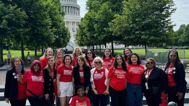 A total of 17 Moms Demand Action and Students Demand Action volunteers, all wearing red branded shirts, pose for a photo during Advocacy Day on Capitol Hill. They stand in front of a staircase with shade trees on either side of the path behind them; the Capitol building is visible between the trees in the background.